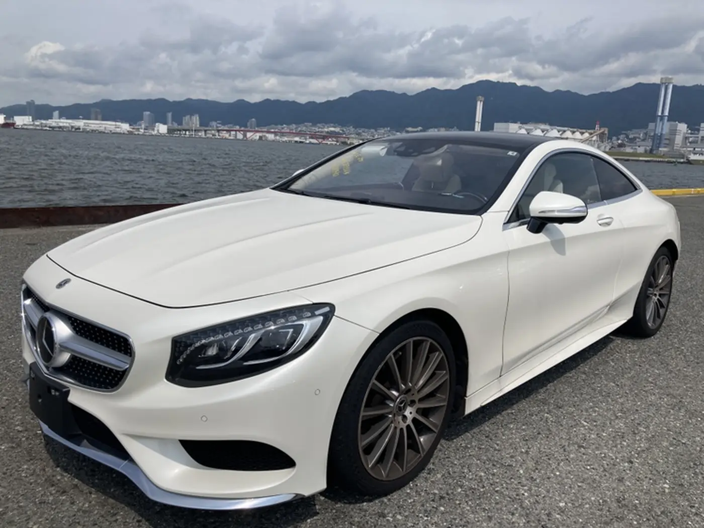 Biały perłowy Mercedes klasy S Coupe widziany z przodu na nabrzeżu, z morzem i górami w tle