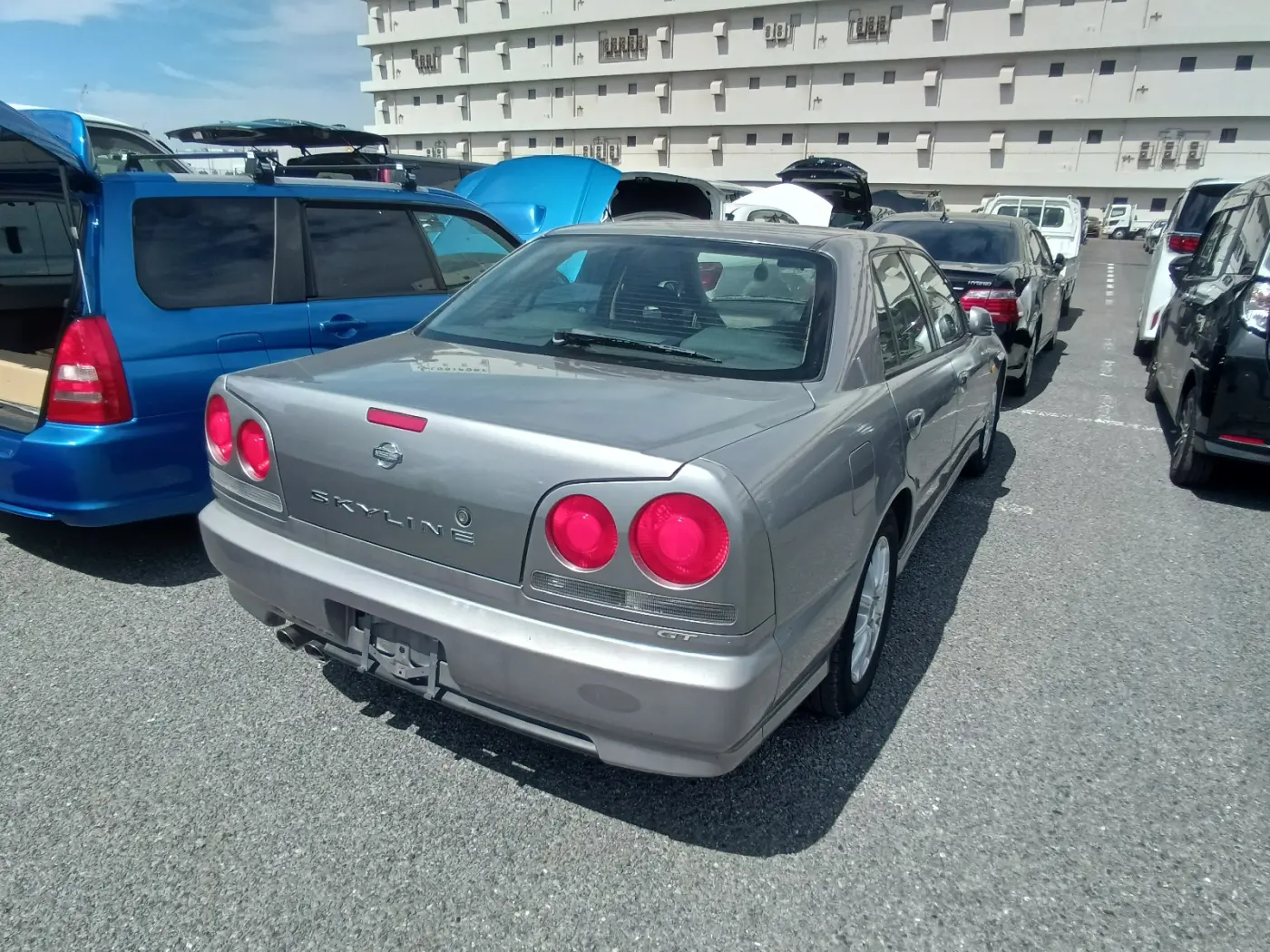 Szary Nissan Skyline GT sedan widziany z tyłu, z okrągłymi czerwonymi lampami, na parkingu aukcyjnym wśród innych aut