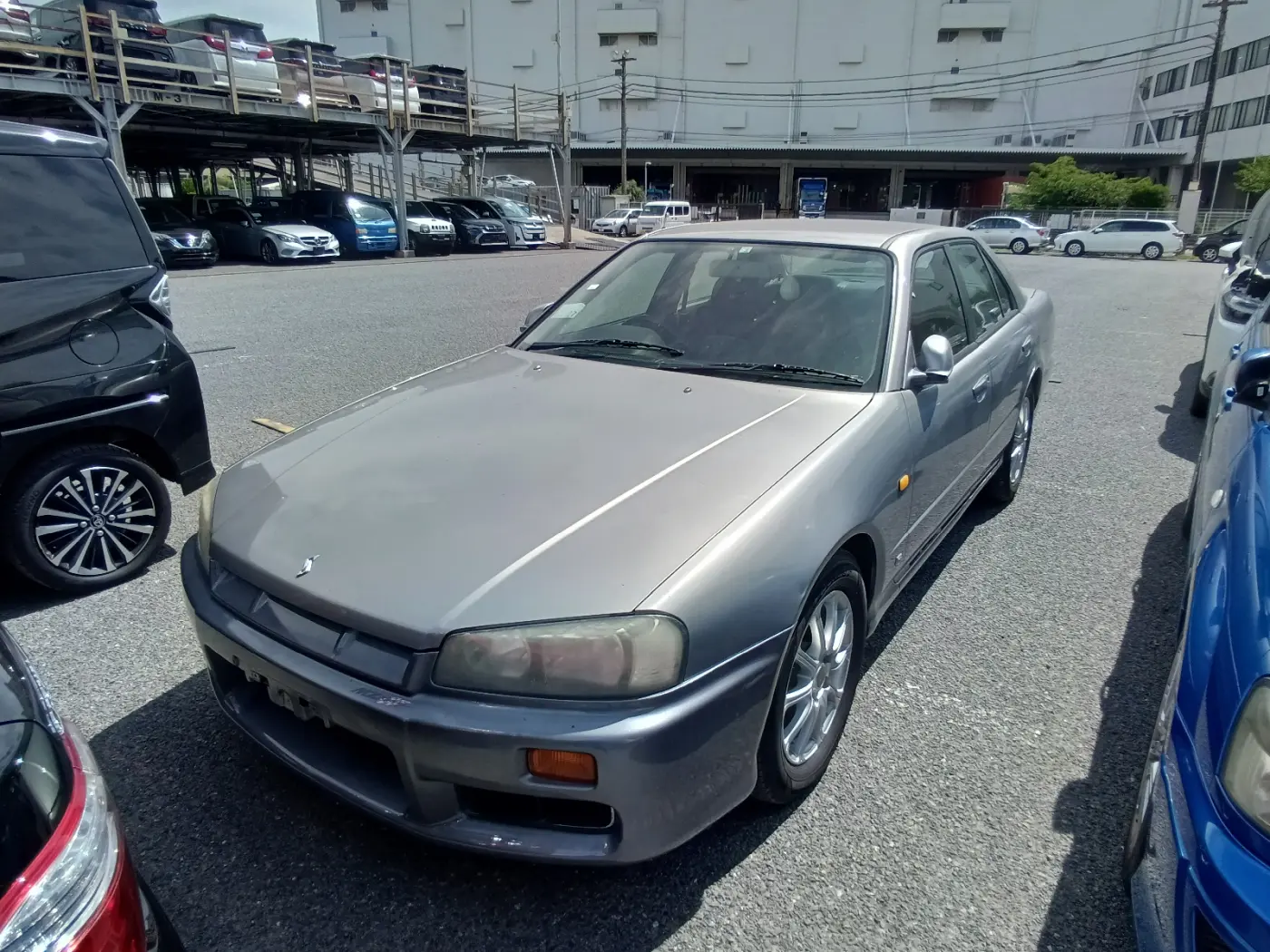 Szary Nissan Skyline sedan widziany z przodu, z zażółconymi reflektorami, na parkingu aukcyjnym wśród innych aut